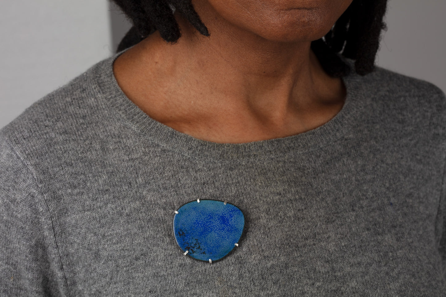 Pebble shaped handmade brooch with shades of dark and light blue and black. Claw set in a silver frame. Worn by a black woman wearing a dark grey jumper.