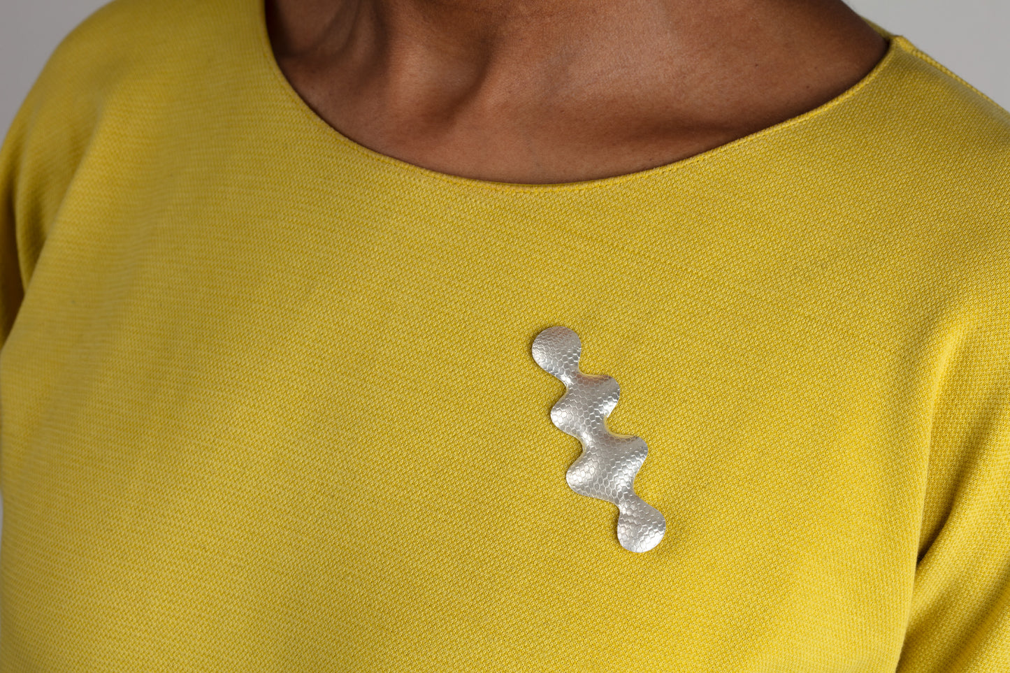 Wavy shaped silver brooch with a textured pattern worn by a woman with a yellow top.