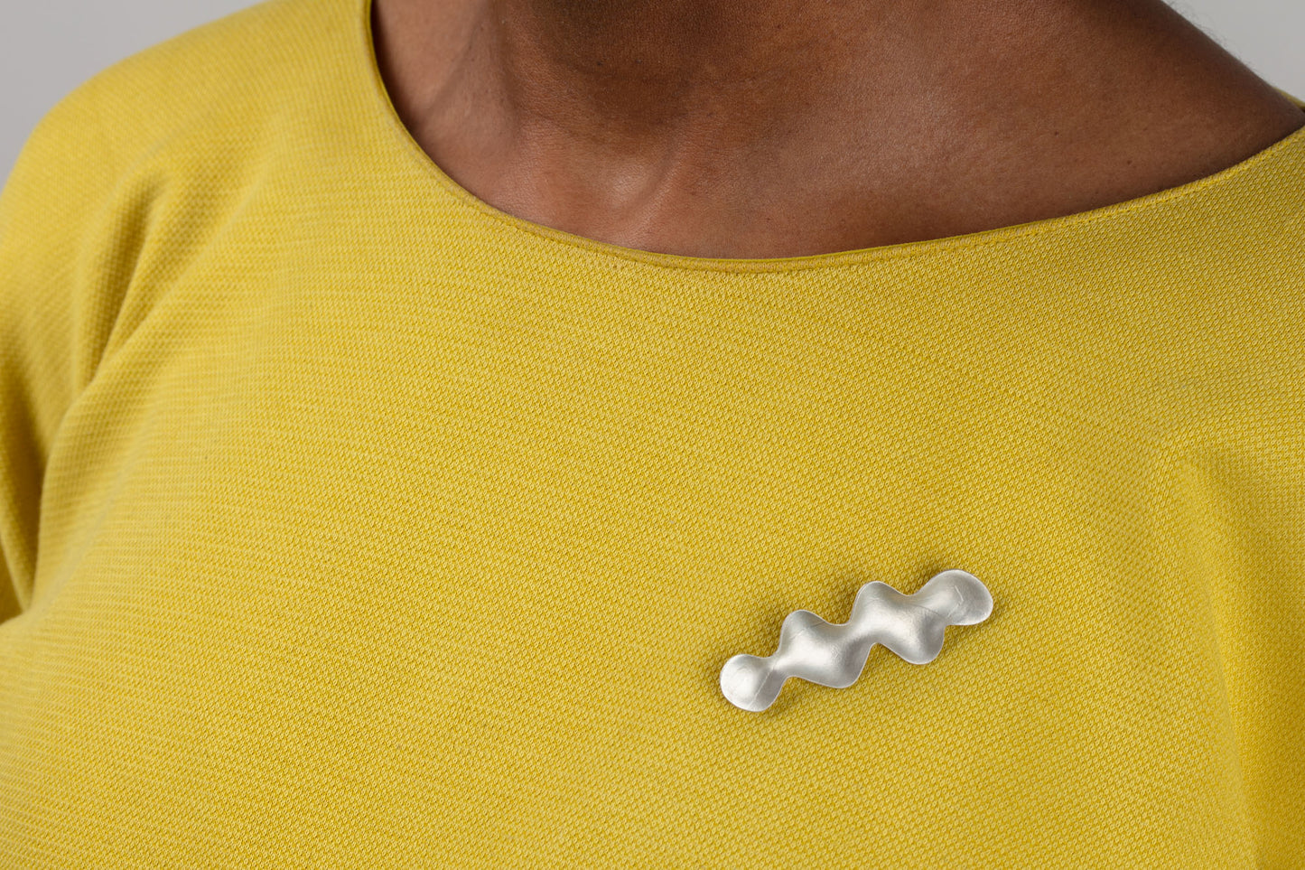Small wave-shaped silver brooch with subtle texture, worn on a yellow top.