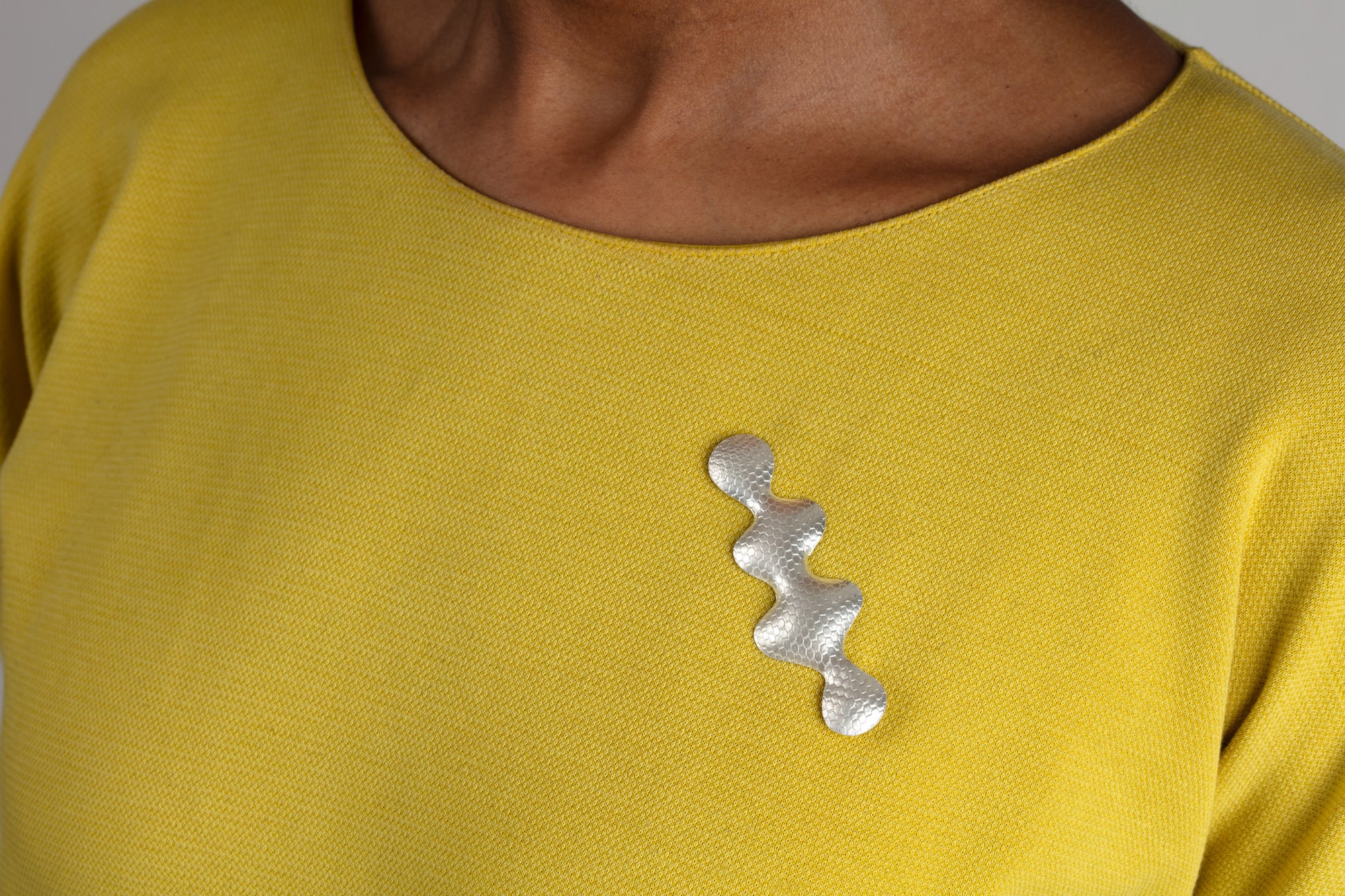 Wavy shaped silver brooch with a textured pattern worn by a woman with a yellow top.