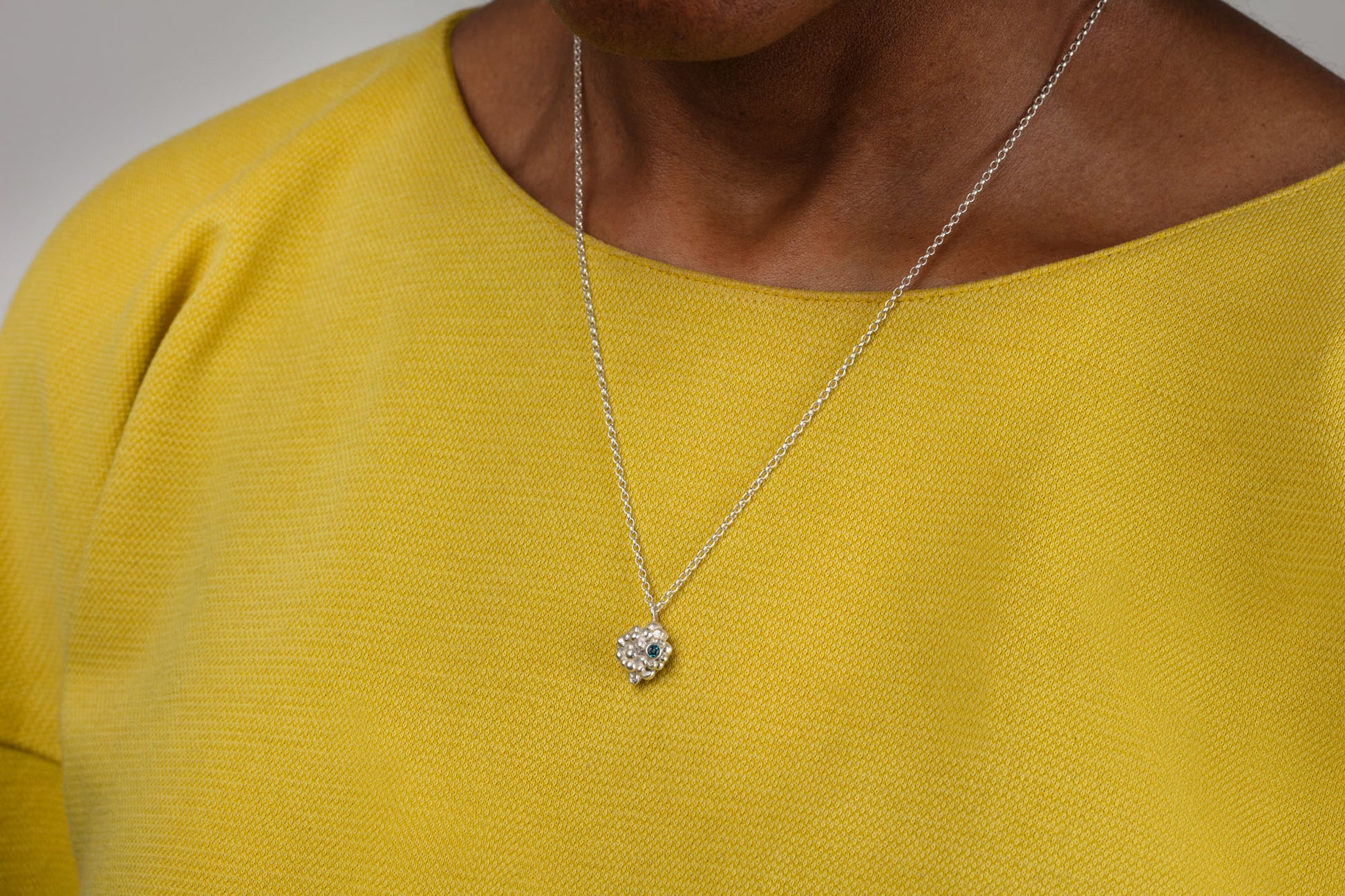 Small silver raspberry shaped pendant made up of lots of little silver balls with a small light blue stone.  The pendant hangs on a silver chain.  Worn by a person with a mustard yellow top.