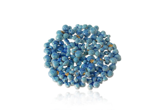 Circular brooch made up of lots of little balls. Each silver granule has been covered in enamel in shades of blue from sky to royal blue.  Some of the ball have small pieces of gold foil that has been added.  The background is white.